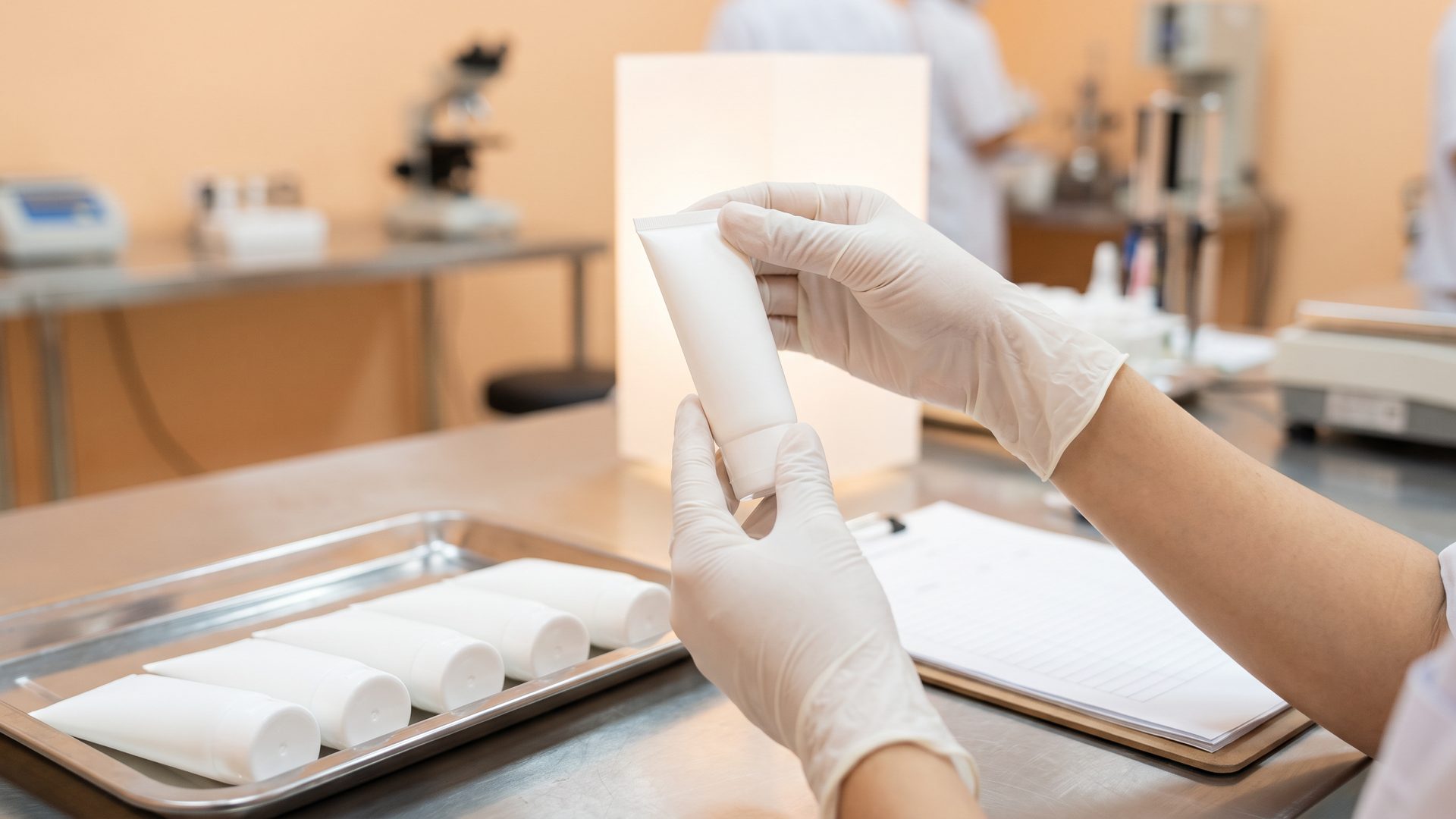 gloved inspector checking a cosmetic tube at a light table with a tray of tubes and a checklist
